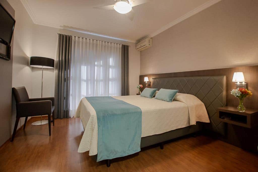 quarto da Pousada Vovó Carolina em Gramado com uma cama de casal decorada com lençol e travesseiros azuis claros. No canto esquerdo do quarto há uma cadeira de courino e uma televisão presa à parede.