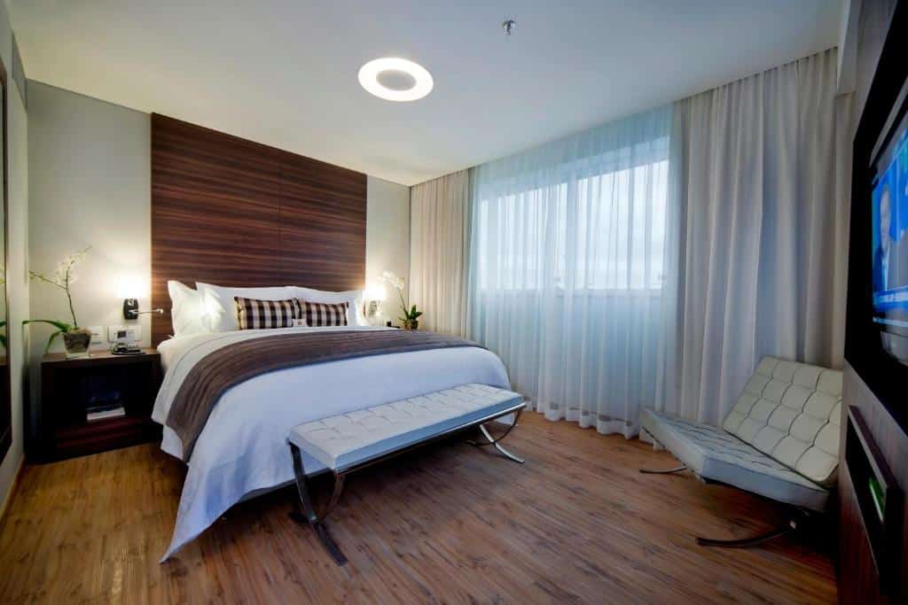 Quarto no Quality Hotel Vitória com cama de casal, recamier, sofá, mesinha com vaso de flores e uma janela com cortina, para representar hotéis em Vitória.