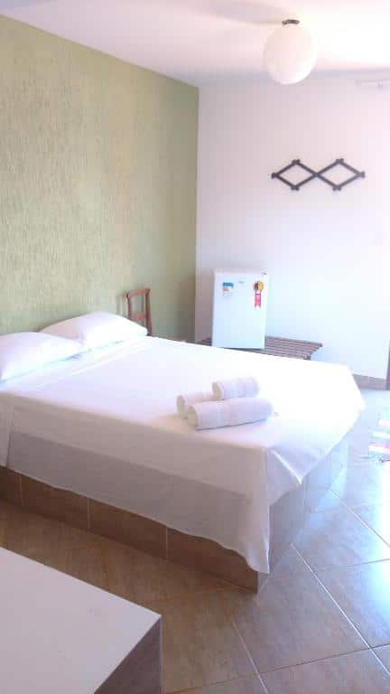 quarto da sinhá cipó com uma cama de casal com lençóis e toalhas brancos, um frigobar e uma cadeira de madeira ao lado da cama.