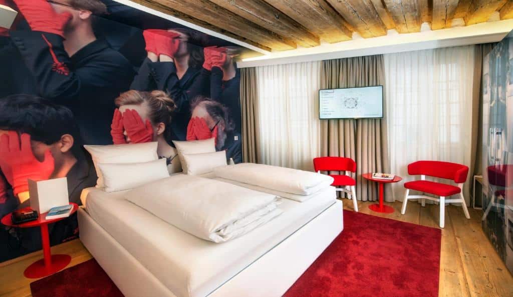 Quarto do hotel com uma cama de casal branca, parede com imagens de várias pessoas com a mão no rosto, duas mesinhas redondas e duas cadeiras vermelhas, tapete vermelho e uma tv na parede com cortina.
