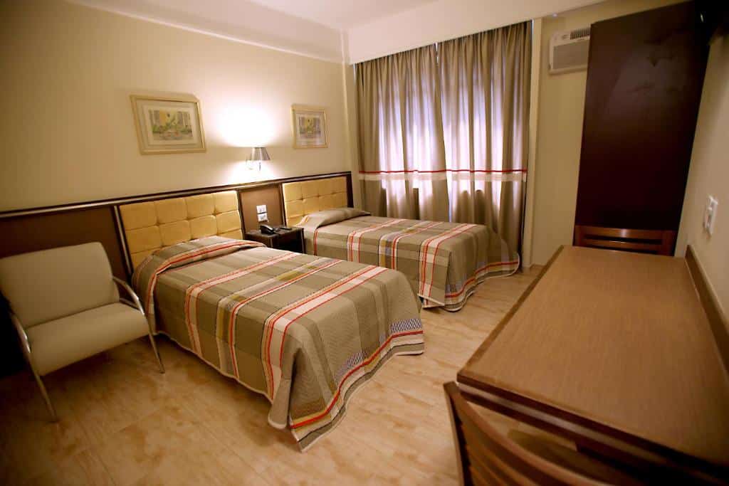 Quarto do hotel com duas camas de solteiro, uma poltrona, uma mesa com cadeiras, dois quadros na parede e uma janela com cortina.
