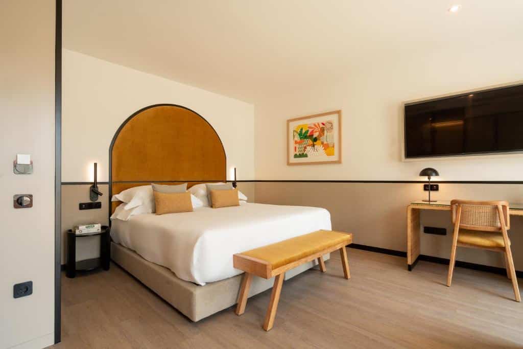 Quarto do The Editory Garden Porto Hotel com cama de casal do lado esquerdo, um banco de madeira no pé da cama e do lado esquerdo da cama, uma mesa de trabalho com uma TV. Representa hotéis de luxo no Porto.