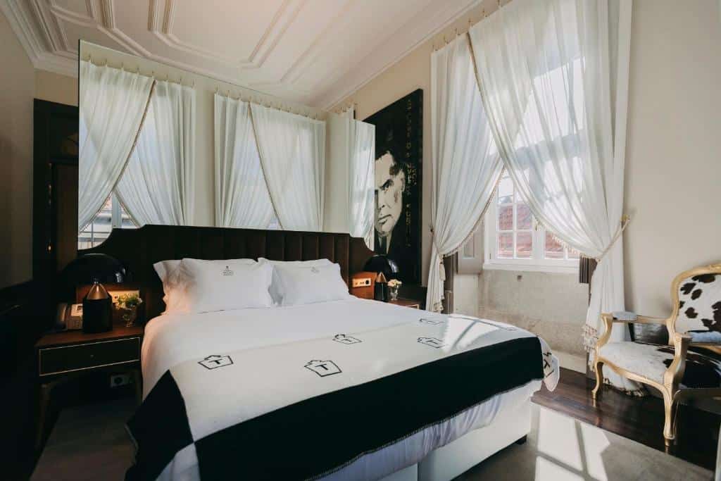 Quarto do Torel Palace Porto com cama de casal do lado esquerdo, duas cômodas do lado da cama com luminária e uma cadeira do lado a frente da cama. Representa hotéis de luxo no Porto.