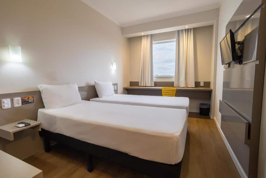 Quarto com duas camas, televisão e uma mesa com cadeira. Acima dela há uma janela com cortina. Foto para ilustrar post sobre hotéis em Campo Grande.