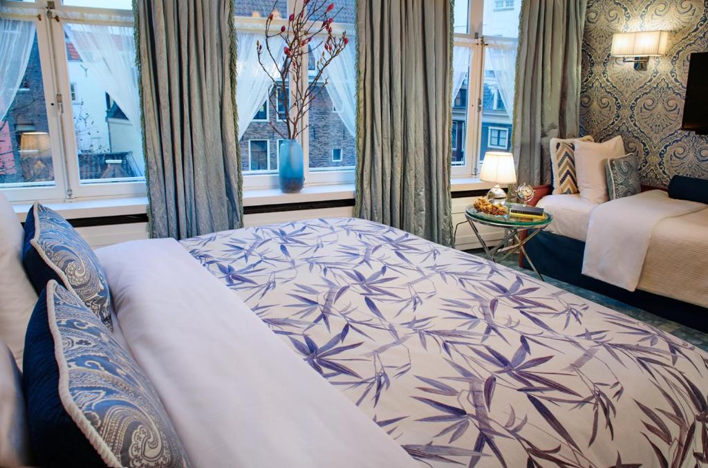 Quarto triplo do Hotel Estheréa, de 22 m², com cama de casal com jogo de cama branco e azul, dois travesseiros em cima no mesmo tom, uma mesinha a frente, ao lado de uma cama de solteiro, e no lado esquerdo tem janelas com amplas com vista para um prédio histórico e algumas cortinas