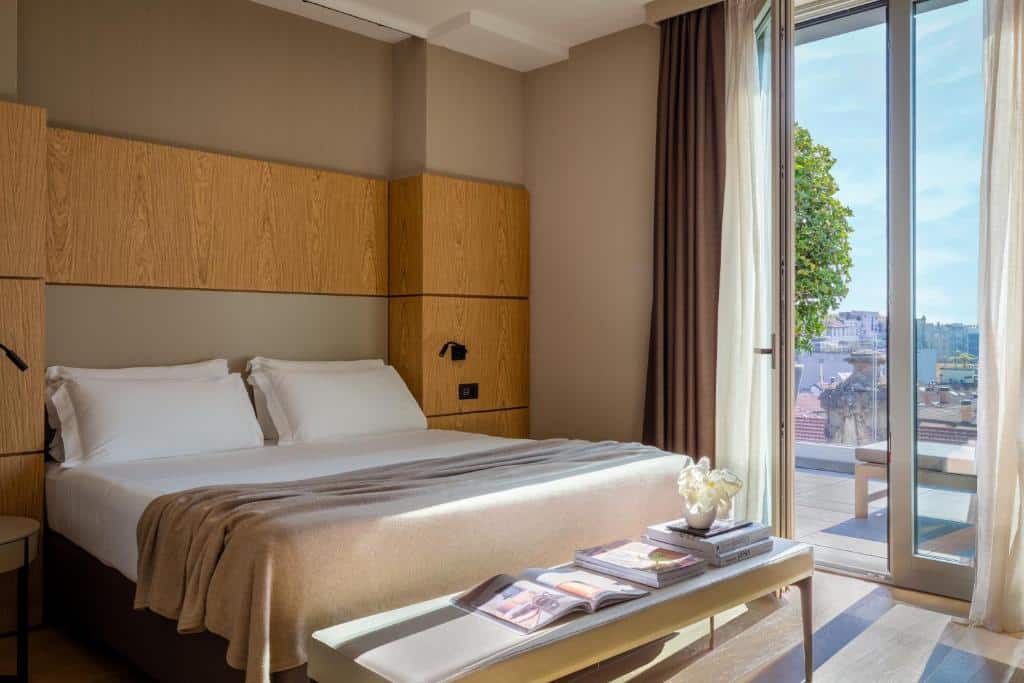 Quarto do Worldhotel Cristoforo Colombo com uma sacada com vista para a cidade, uma cama de casal, um pequeno sofá na beira da cama com livros e um vaso de flor