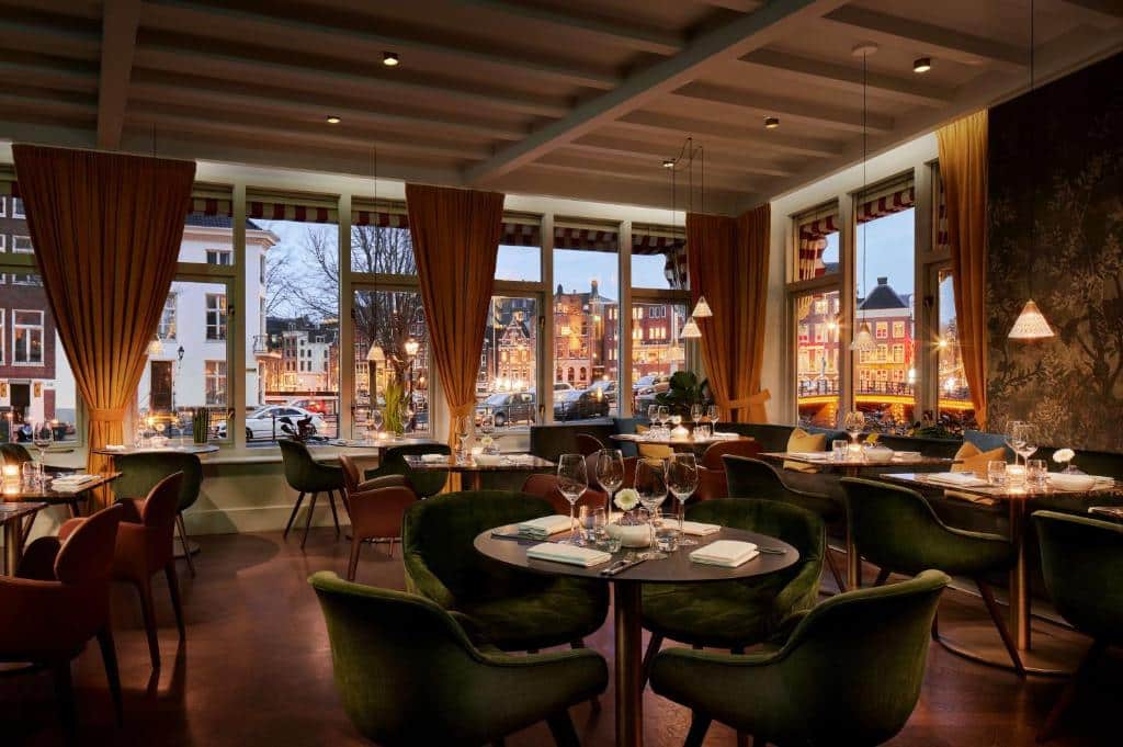 Restaurante do Tivoli Doelen Amsterdam Hotel. com mesas redondas e cadeiras verdes estofadas, e em todas as paredes há janelas amplas de vidro com vista da cidade