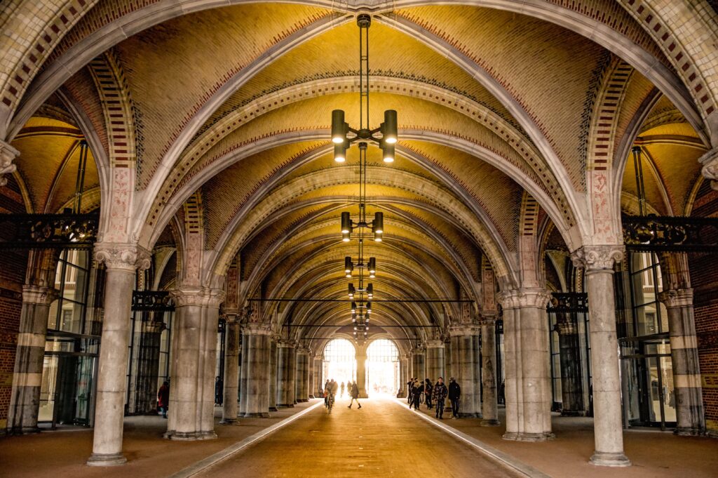 Interior do Rijksmuseum, sendo o local histórico, com o teto com arcos ovais, lustres e várias colunas. Há pessoas andando e nos lados direito e esquerdo tem portas pretas de vridro.
