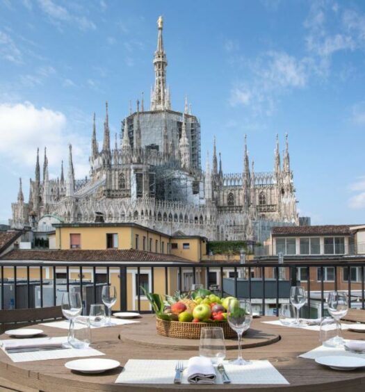 Varanda do hotel Rosa Grand Milano – Starhotels Collezione com uma mesa com pratos, talheres e guardanapos em pano, de frente para a varanda, é possível ver o topo da Catedral de Milão, para representar hotéis perto do Duomo em Milão