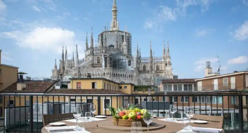 Varanda do hotel Rosa Grand Milano – Starhotels Collezione com uma mesa com pratos, talheres e guardanapos em pano, de frente para a varanda, é possível ver o topo da Catedral de Milão, para representar hotéis perto do Duomo em Milão