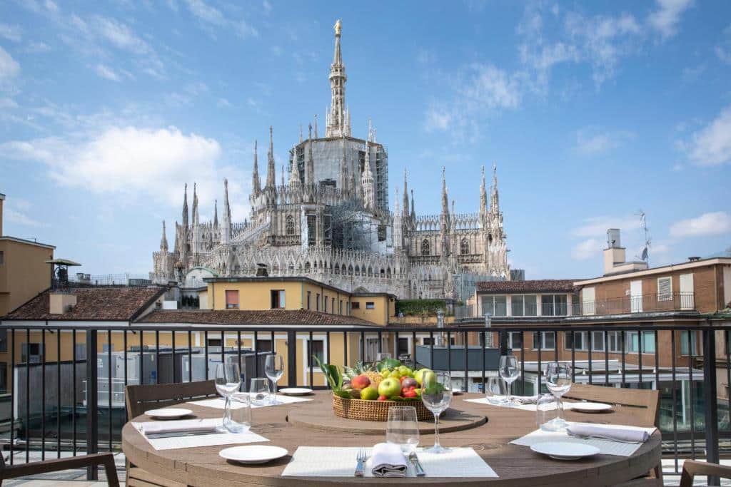Varanda do hotel Rosa Grand Milano – Starhotels Collezione com uma mesa com pratos, talheres e guardanapos em pano, de frente para a varanda, é possível ver o topo da Catedral de Milão, para representar hotéis perto do Duomo em Milão