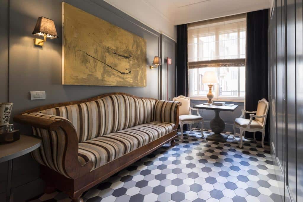 área de estar da Casa Montani com chão desenhado, sofá listrado, quadro na parede, mesinha com poltronas e uma janela ampla com cortinas, tudo em estilo vintage