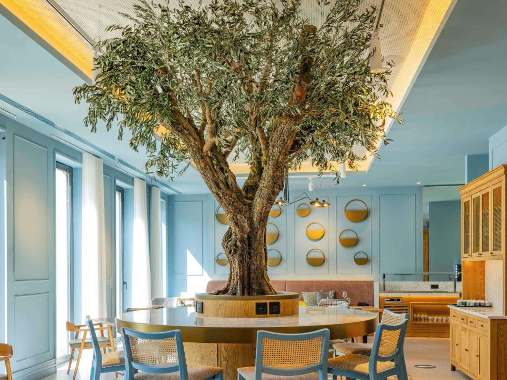 Sala de refeição do Mercure Porto Centro Aliados com cadeiras e mesas no ambiente e no meio uma bela árvore. Representa onde ficar no Porto.