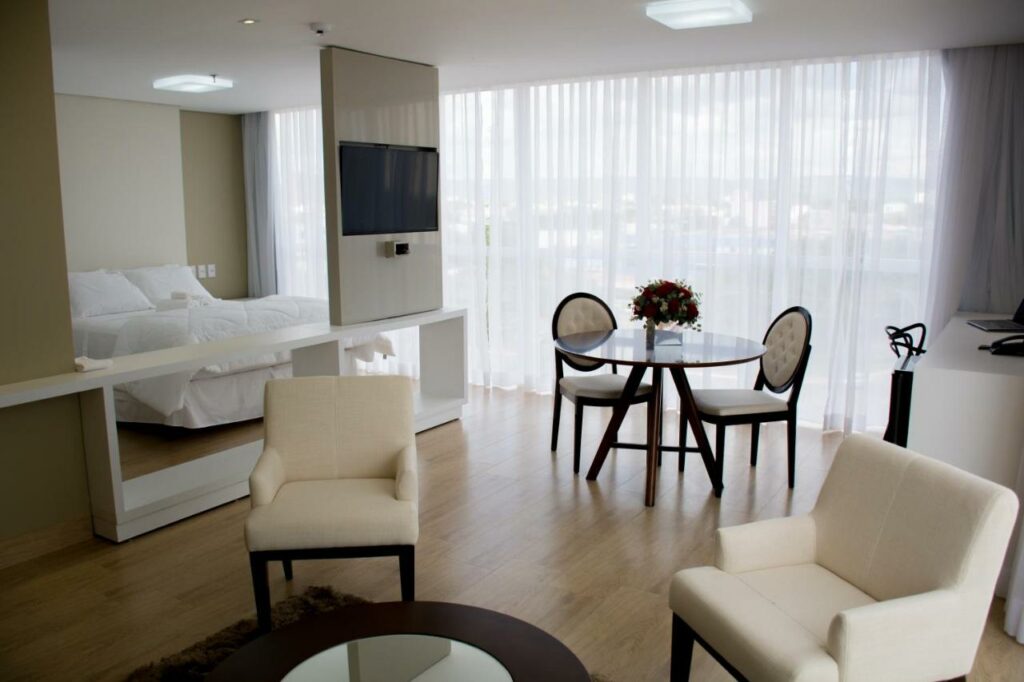 Quarto do Select Hotel com cama de casal, televisão, poltronas, mesas e cadeiras e janela com cortina branca. Foto para ilustrar post de hotéis em Palmas.