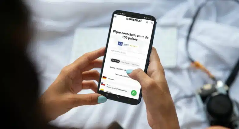 Mulher, sentada na cama, aparecendo na imagem metade da sua cabeça embaçada, segurando com as duas mãos um celular com a tela mostrando o site da Sim Premium com opções de planos do chip e eSIM Premim.