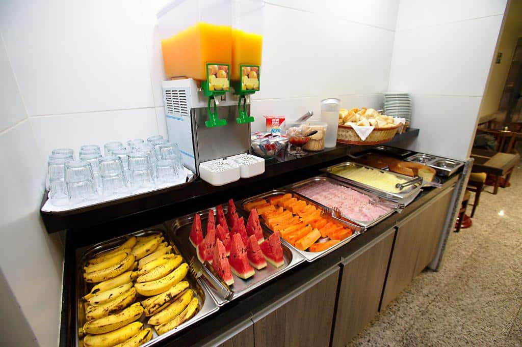 Café da manhã exposto com algumas frutas, como banana, melancia e mamão. Alguns frios, sucos, pães e copos de vidro, ilustrando post Hotéis perto da 25 de Março em SP.