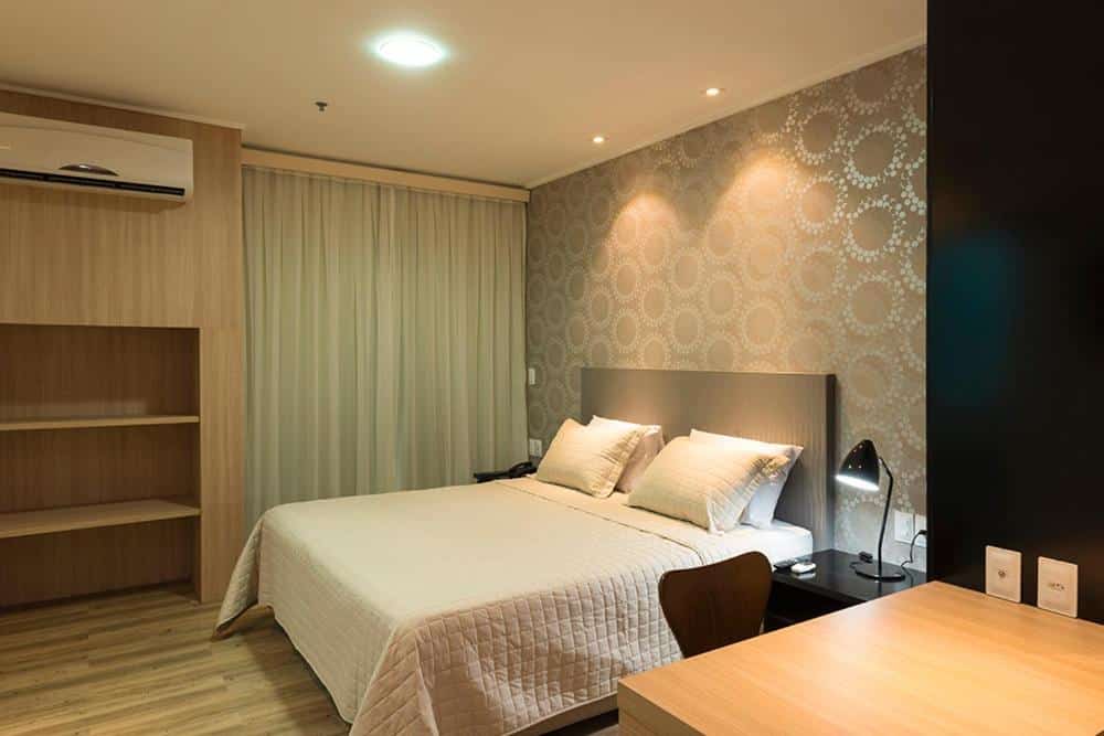 Quarto moderno com papel de parede decorativo, decoração em tons claros, cama de casal com colcha branca e cortinas claras. Imagem para ilustrar o post hotéis em Teresina.