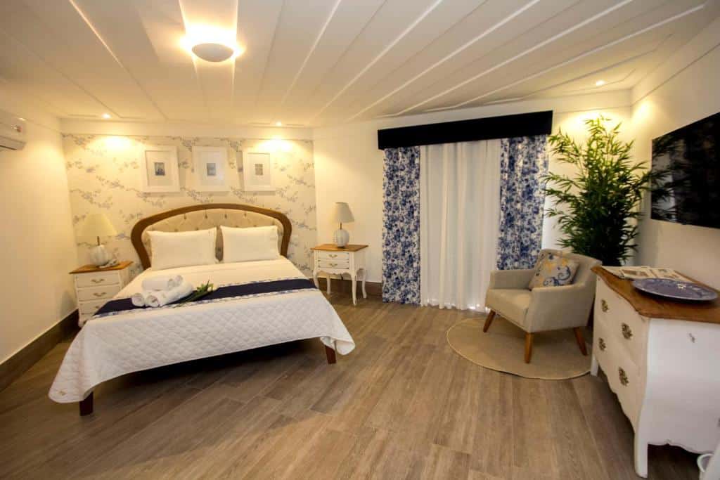 Grande quarto de hotel com cama de casal, decoração em tons de azul e branco, abajour, poltrona, mesinhas, cômoda e televisão. Imagem para ilustar o post hotéis em Cabo Frio.