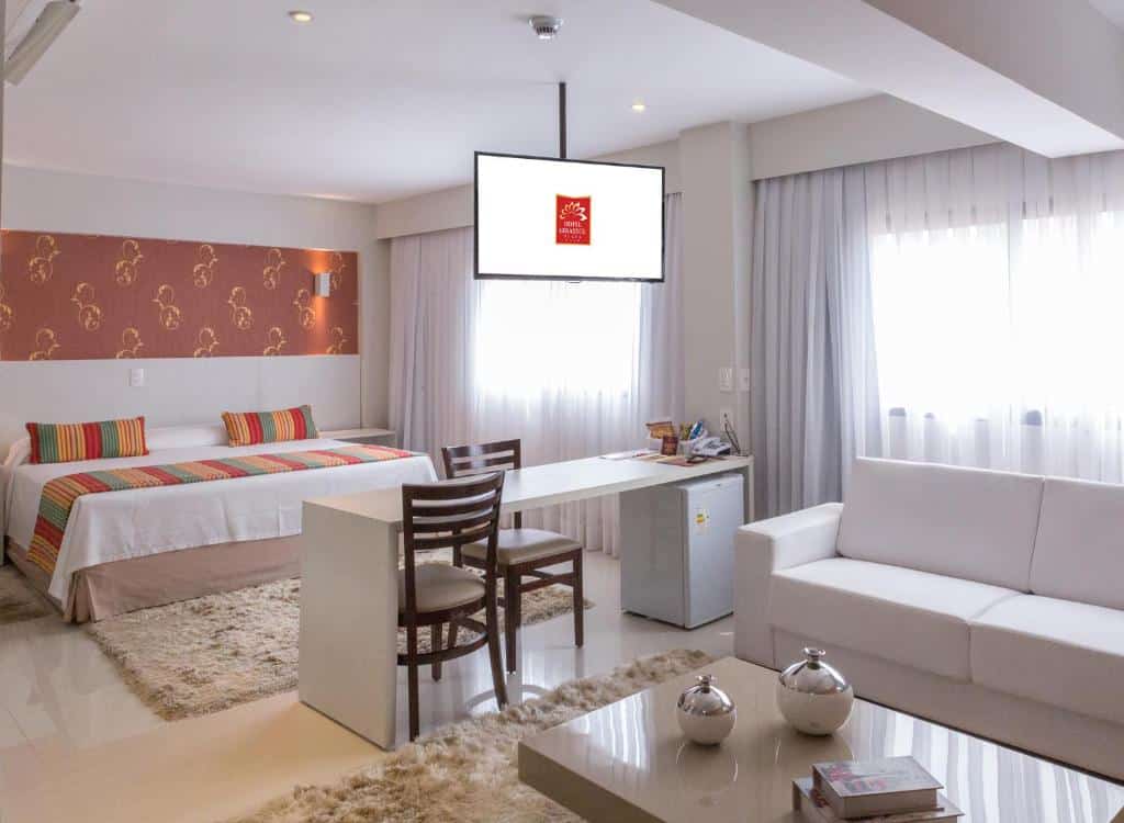 Quarto do Hotel Girassol Plaza com uma cama de casal, mesa com cadeiras, sofá branco, televisão e janelas grandes com cortinas brancas. Foto para ilustrar post de hotéis em Palmas.