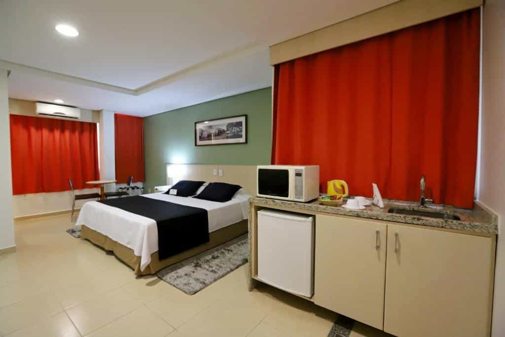 Quarto com cama de casal no lado esquerdo e cozinha compacta no lado direito, cortinas vermelhas, parede em tom de verde e quadro decorativo. Imagem para ilustrar o post hotéis em Goiânia.