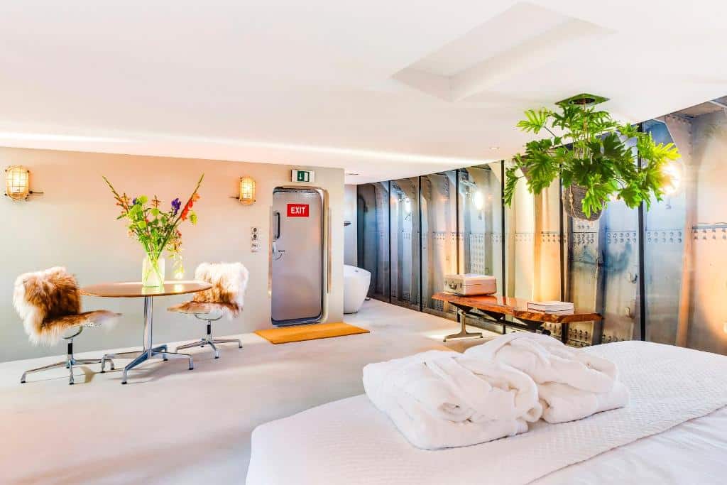 Suite King Deluxe, de 60 m², do Lightship Amsterdam, com cama de casal com roupões dobrados em cima, uma mesa com duas cadeiras e um vaso acima da mesa e , ao fundo, uma porta escrito "EXIT" com uma banheira ao lado.
