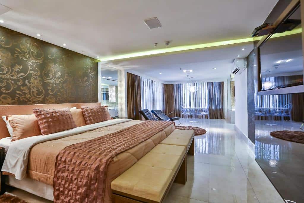 Quarto espaçoso com grande cama de casal com colchas rose, papel de parede a cima da cama e mesa no fundo do quarto com cortinas atrás. Imagem para ilustrar o post hotéis em Goiânia.