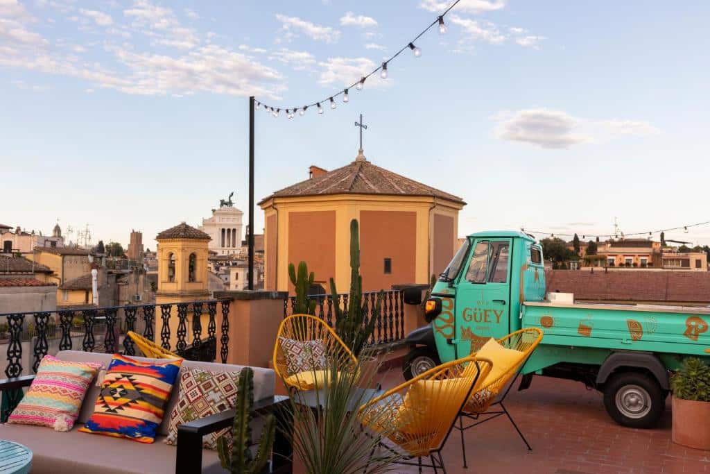 terraço com vista da cidade do Chapter Roma com mesa com cadeiras amarelas e sofá neutro com almofadas coloridas, há, ainda uma kombi descontruída, meio lambreta, verde