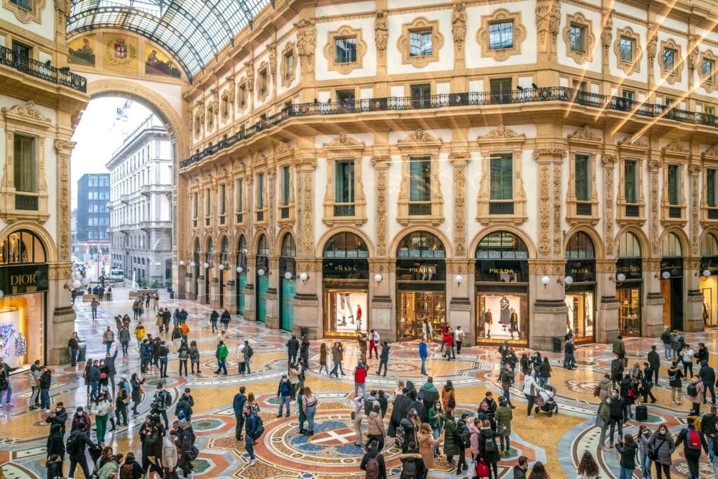 Parte interna da Galeria Vittorio Emanuele II, com uma arquitetura renascentista, piso pintado, teto de vidro, lojas de marcas e muitas pessoas nadando no local