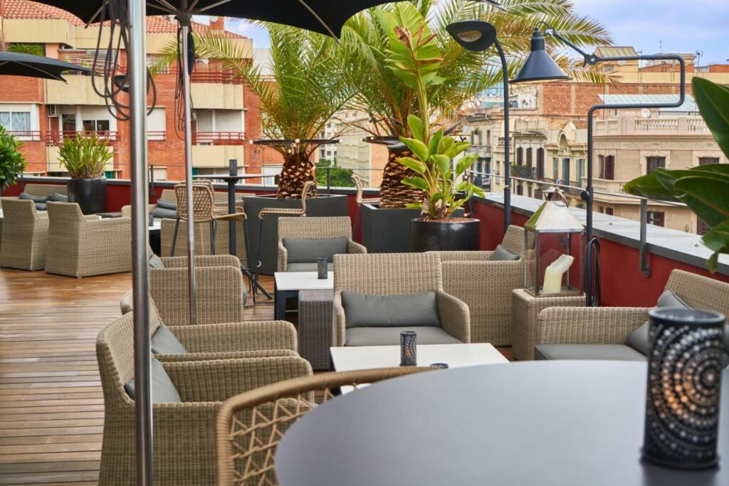 Terraço do Hotel Villa Emilia. Diversas cadeiras e mesas estão espalhadas pelo local, com abajures e vasos de plantas intercalados entre si. Há vista para a cidade de Barcelona.