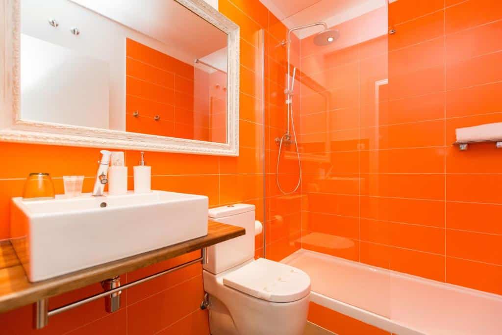 Banheiro do Violeta Boutique. Com azulejos laranja, o local tem uma privada entre a banheira com box e chuveiro e a pia com amenidades de banho e espelho.
