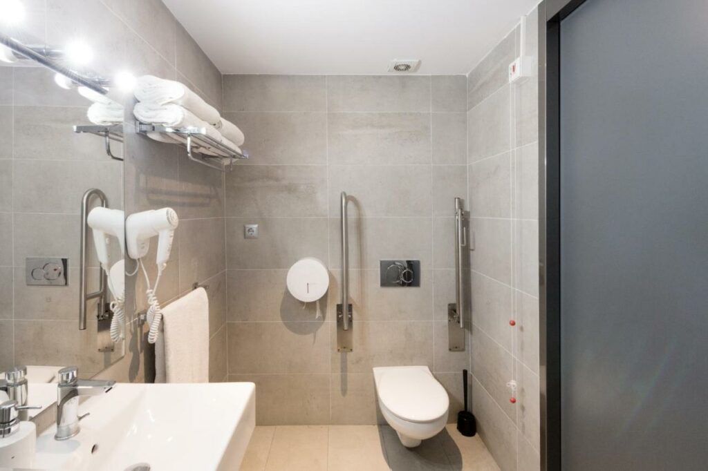Banheiro adaptado do Yeah Barcelona Hostel, uma das recomendações de hostels em Barcelona com acessibilidade. Há barras de apoio dos dois lados da privada. A pia branca tem um espelho na parede acima e um secador de cabelos ao lado. Há suportes com toalhas brancas ao lado da pia.