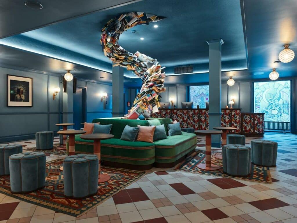 Área comum do 25hours Hotel Indre By com vários sofás, puffs, balcão de atendimento e uma escultura feita de livros bem no meio.