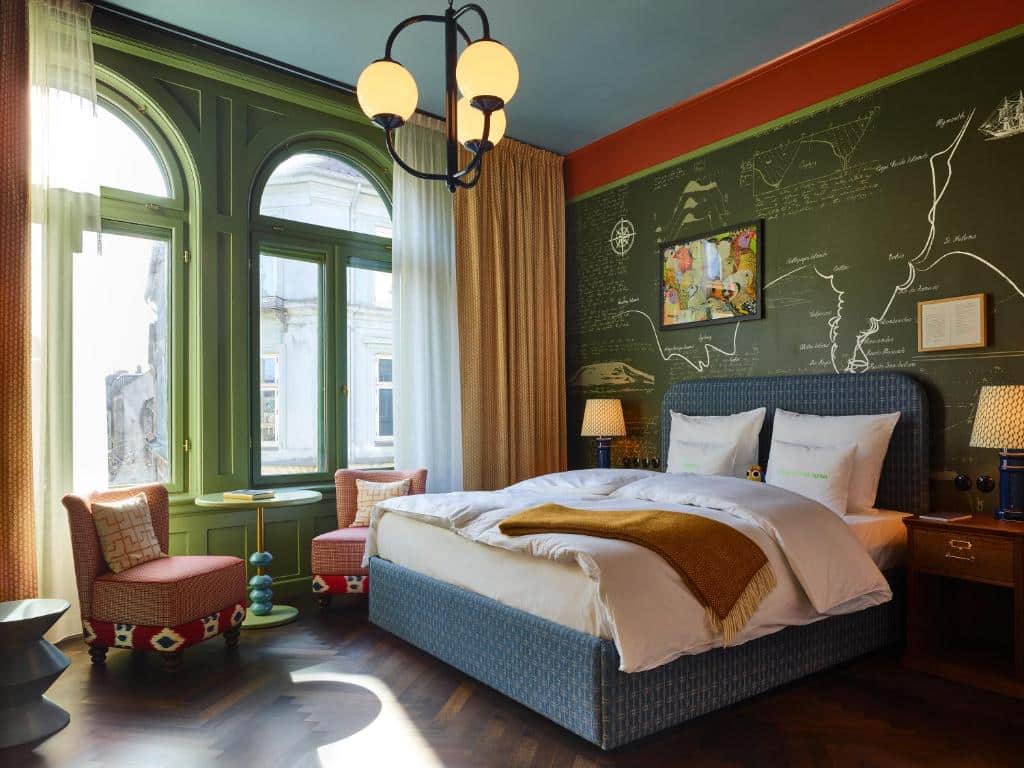 Quarto do 25hous Hotel Indre By com uma cama de casal, duas mini poltronas, mesinha, abajur e uma parede toda desenhada. O quarto é todo colorido e há duas janelas com vista para a ciadade. Foto para ilustrar post hotéis em Copenhague.