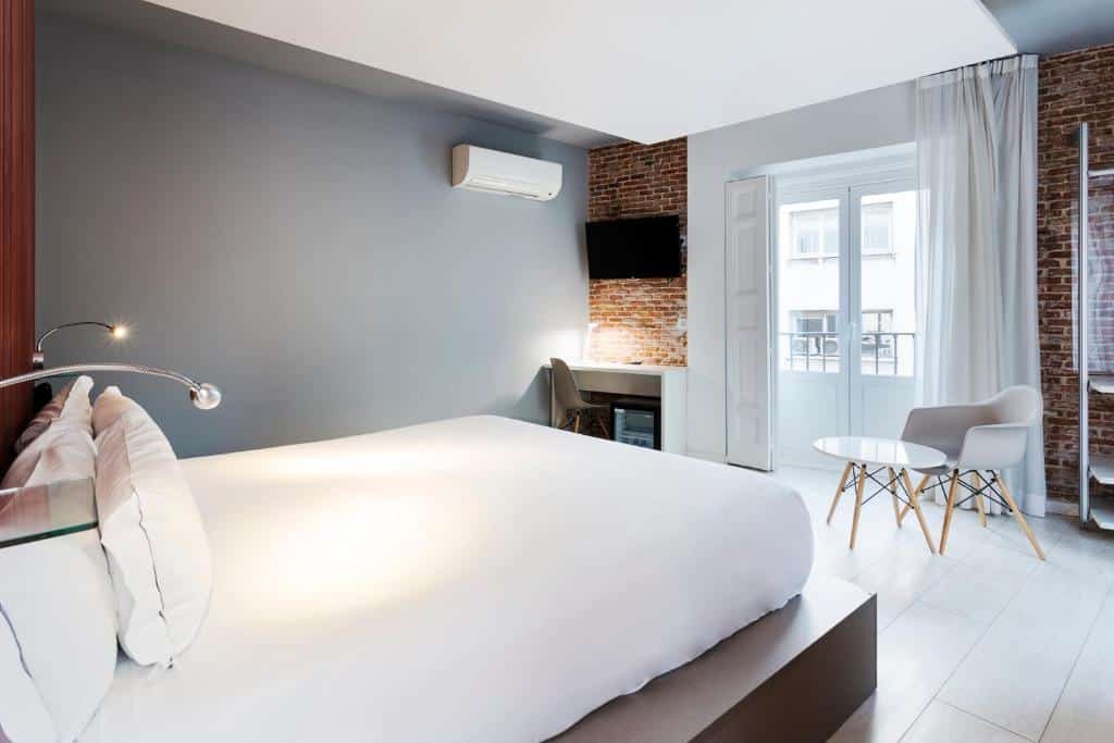 Quarto do B&B HOTEL Madrid Centro Fuencarral 52, uma das recomendações de hotéis baratos em Madri. Uma cama de casal tem luminárias logo acima e encara uma área de estar com mesa e cadeira em frente a uma janela com cortinas brancas diáfanas. Ao lado está uma mesa de trabalho com cadeira e uma TV na parede acima. O aparelho de ar-condicionado está logo ao lado.