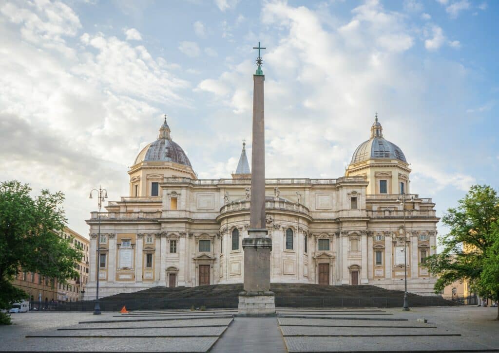 vista da Basílica Santa Maria Maggiore, em Roma, com abóbadas trabalhadas em estilo bem clássico com fachada branca e há um obelisco à frente
