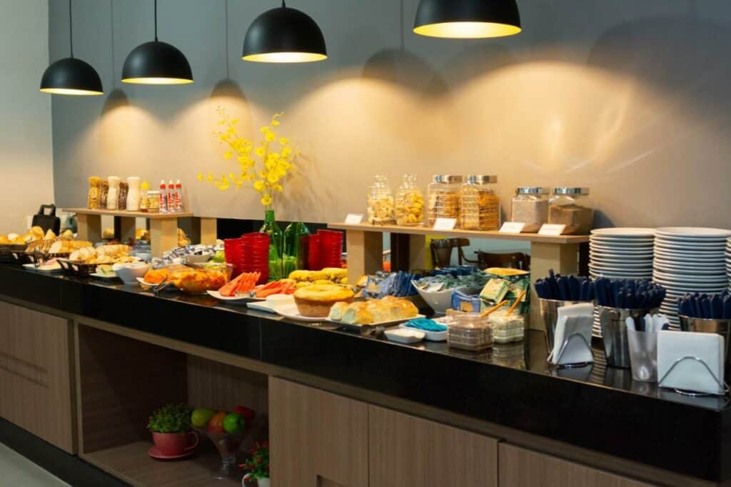 Mesa de café da manhã do Abudi Hotel com algumas frutas, pratos e talheres, pães, bolos e outros tipos de comidas.