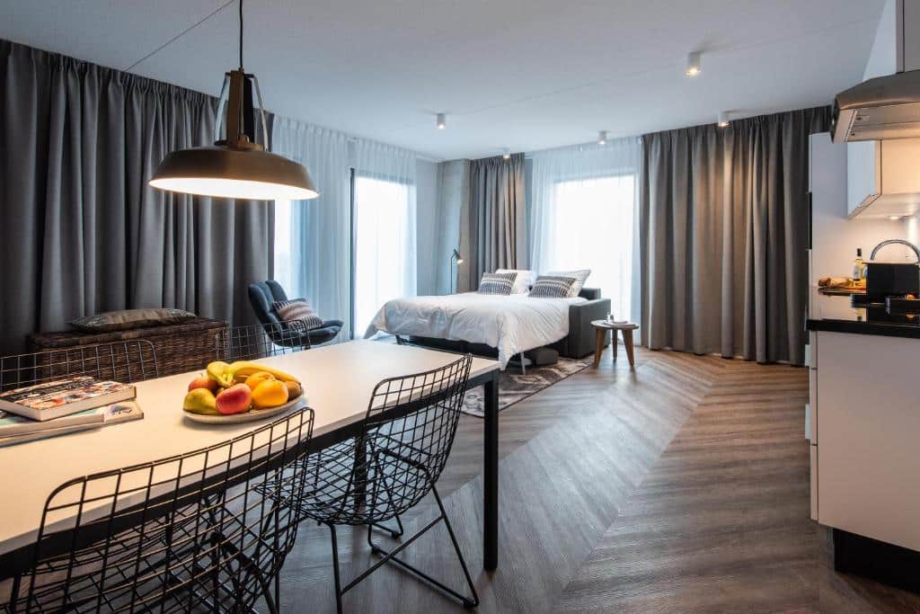 Apartamento de 1 quarto para 4 pessoas do YAYS Amsterdam Docklands, de 55 m², com mesa de 4 cadeiras com uma fruteira em cima com frutas, no lado direito tem uma pia da mini cozinha e no lado esquerdo a cama de casal e uma poltrona. Há bastante espaço entre os móveis e a imagem representa hotéis para família em Amsterdam