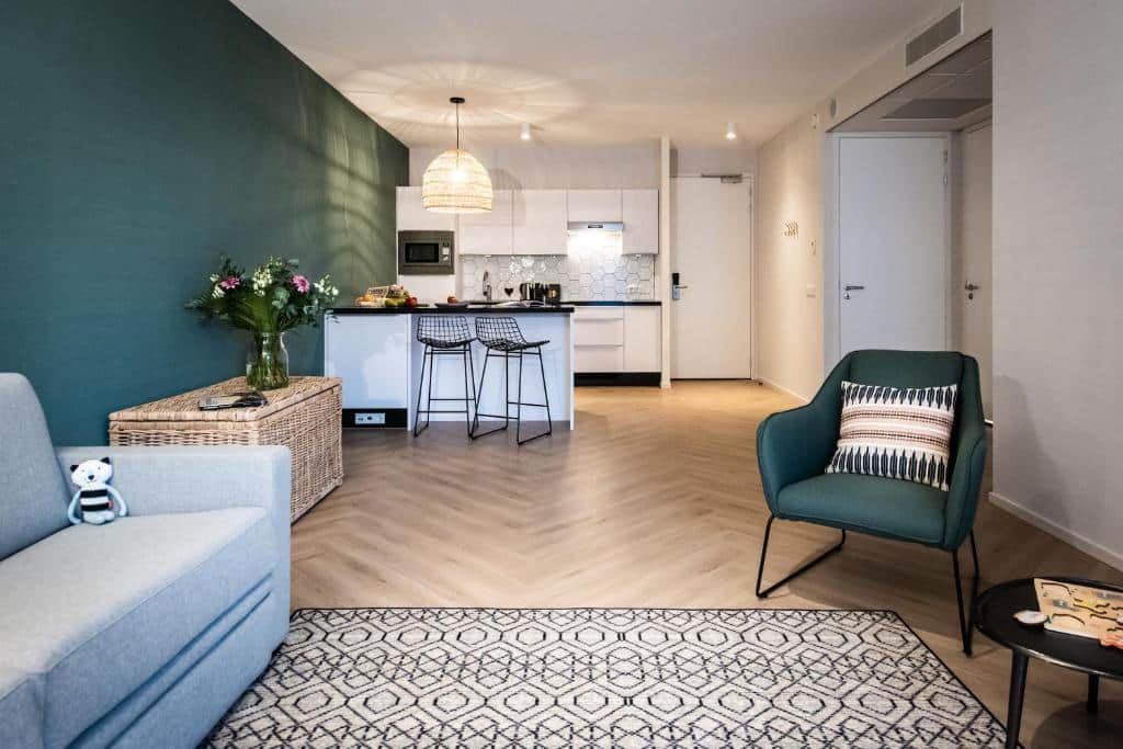 Sala de estar com cozinha compacta de um dos apartamentos do YAYS Amsterdam Docklands, há um sofá com um ursinho em cima, uma poltrona verde, um móvel com vaso de flor em cima e, ao fundo, tem um balcão com bancos a frente e  a cozinha atrás