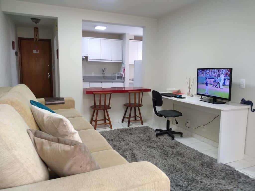 Sala do apartamento com um sofá grande marrom claro, uma tv em cima da mesa branca e ao lado uma cadeira. duas cadeiras de madeira e uma bancada da cozinha, ilustrando post Hotéis em Maringá.