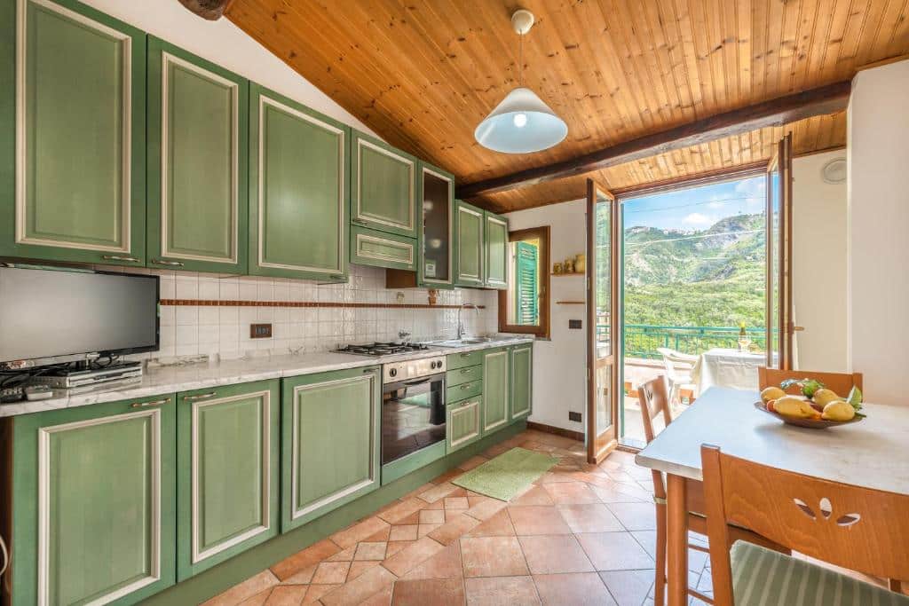 Cozinha com armários verdes, uma TV na bancada, um fogão, uma mesa com três cadeiras de madeira e uma porta grande que dá acesso a varanda, ilustrando post Hotéis em Cinque Terre.