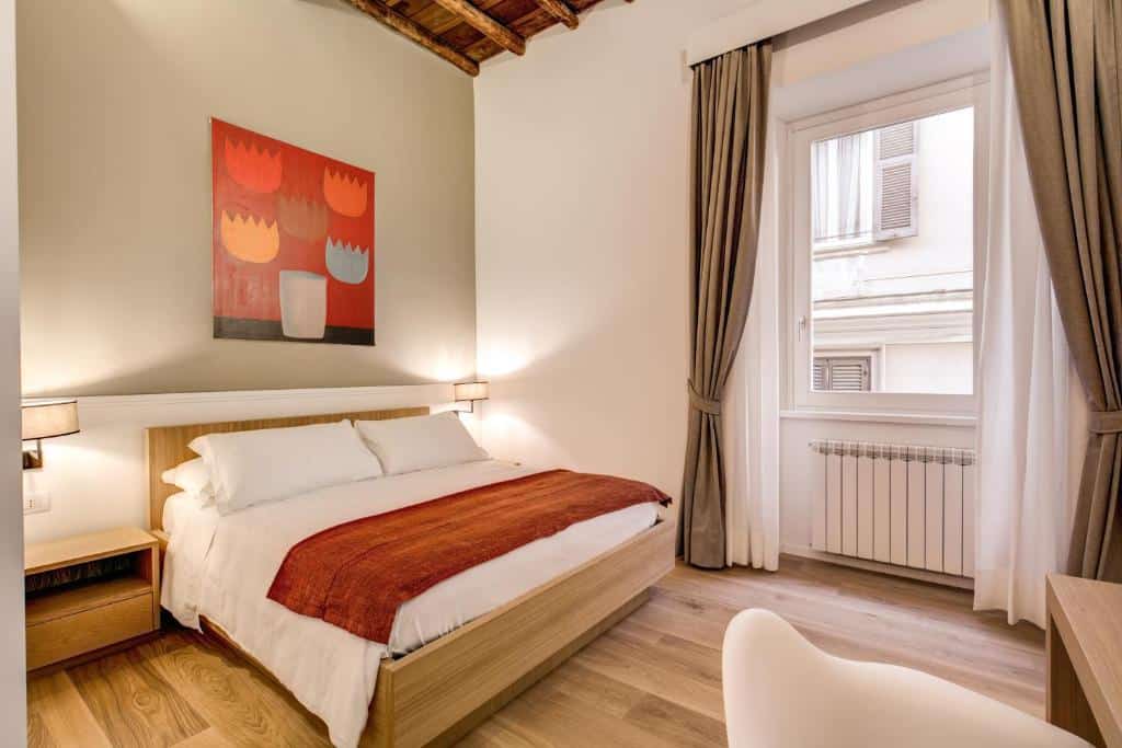 aluguel de temporada no Dopodomani Suite, em Roma, com cama de casal, poltrona branca e mesa à frente e os detalhes em vermelho, com quadro na parede, há luminária e mesinha de ambos os lados, a janela é bem grande com cortinas