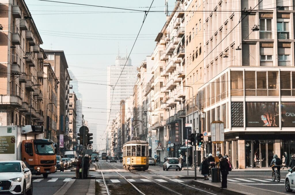 Uma rua de Milão com prédios antigos, pessoas caminhando, há carros e também bondinhos na rua