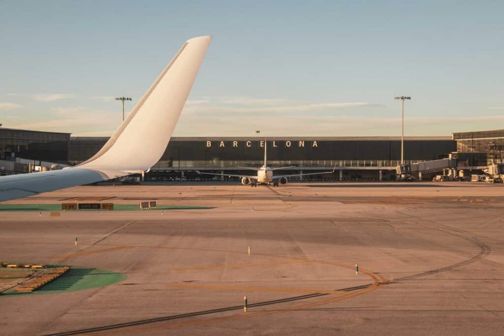 Terminal do aeroporto El Prat para ilustrar o post sobre aluguel de carro no aeroporto de Barcelona. A cauda de um avião está em primeiro plano no canto esquerdo, e ao fundo há outro avião em frente à placa com o nome da cidade na fachada. - Foto: Angela Compagnone via Unsplash