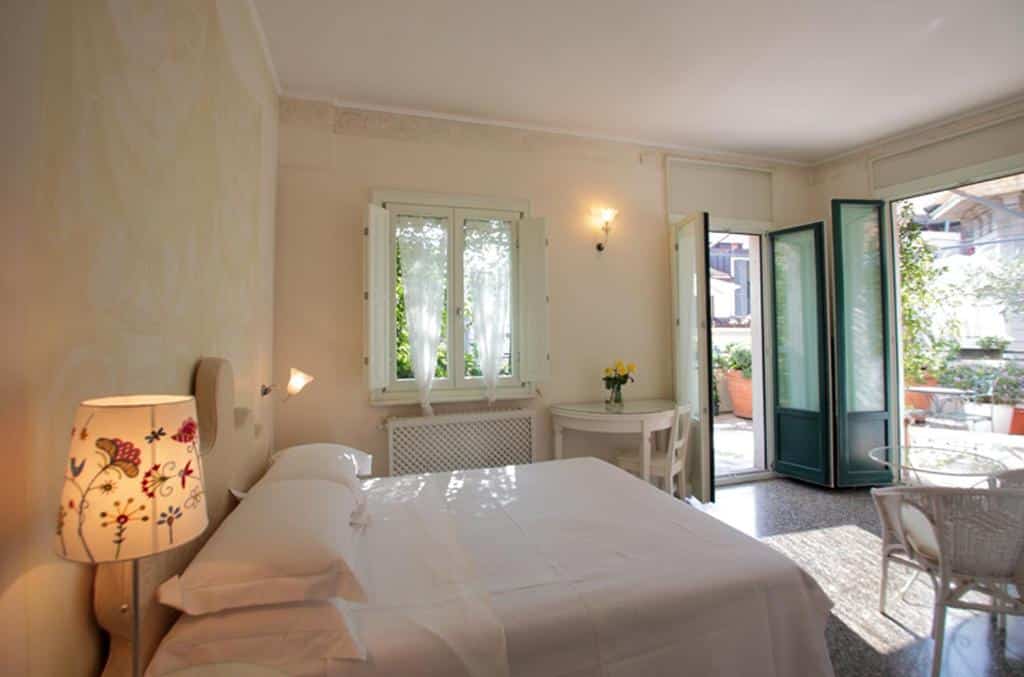 Quarto do Antica Locanda Dei Mercanti  com duas portas que levam pra um terraço com vasos de plantas, o quarto é todo branco com uma cama de casal, uma mesinha redonda com duas cadeiras