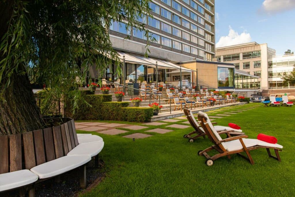 Área externa do Hilton Amsterdam, com um jardim, gramado, espreguiçadeiras e uma área com degraus e cadeiras ao ar livre. Esses degraus dão acesso ao prédio espelhado do hotel