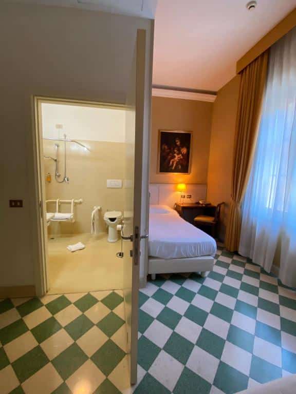 vista do quarto e banheiro do Basilica Hotel, um dos hotéis bem localizados em Roma, com vaso sanitário adaptado, barras de apoio e cadeira de banho