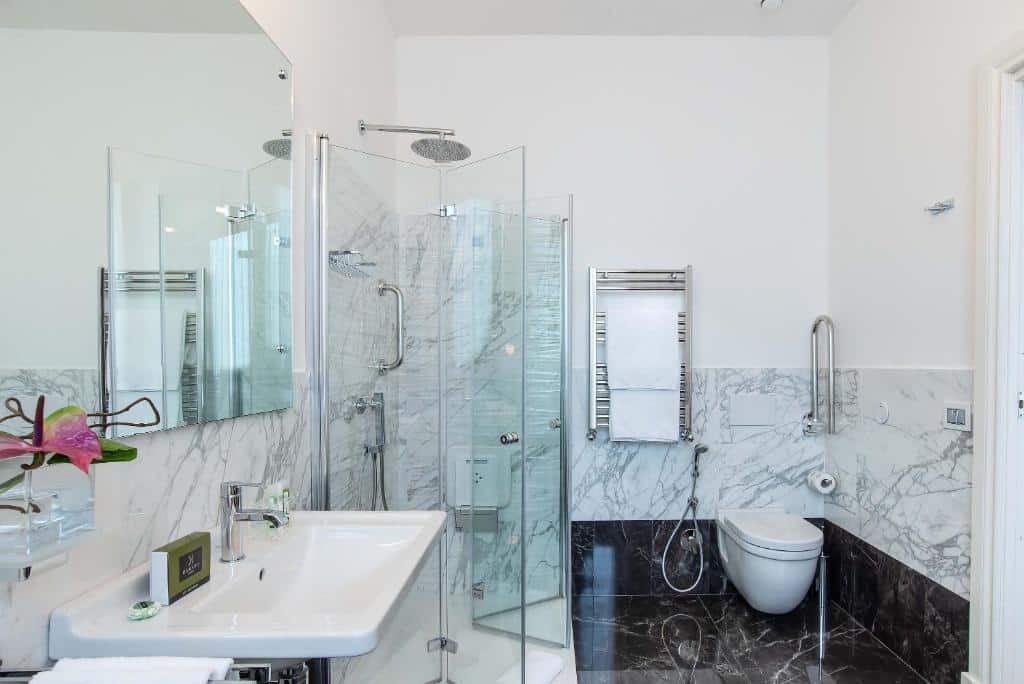 banheiro acessível do Hotel Damaso, um dos hotéis românticos em Roma, com pia com espaço embaixo, vaso sanitário mais alto, box com porta sanfonada, chuveirinho e barras de apoio em todo o ambiente