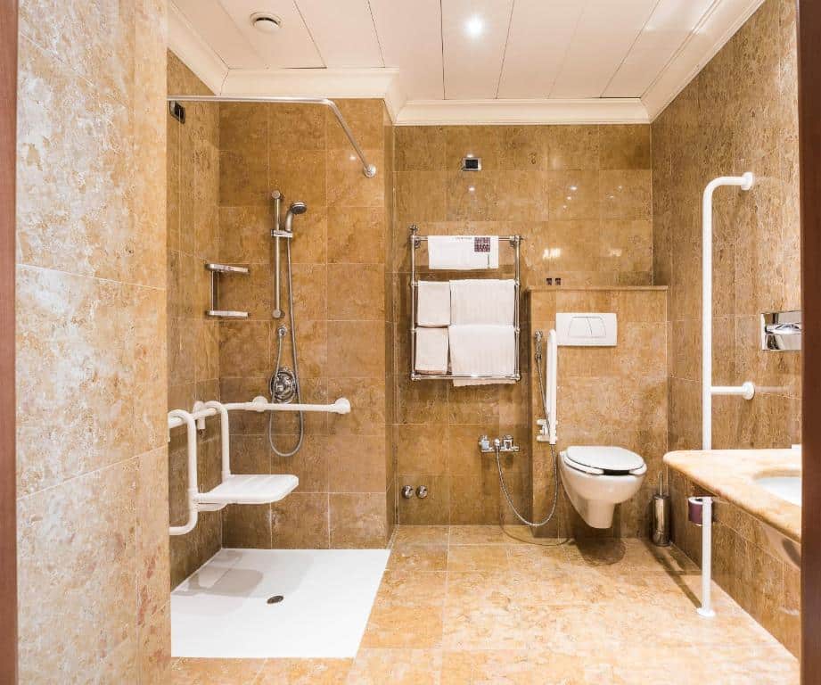 banheiro do Hotel Ponte Sisto, um dos hotéis para brasileiros em Roma, com vaso sanitário mais alto, pia mais baixa com espaço, cadeira de banho no box aberto com chuveirinho e muitas barras de apoio