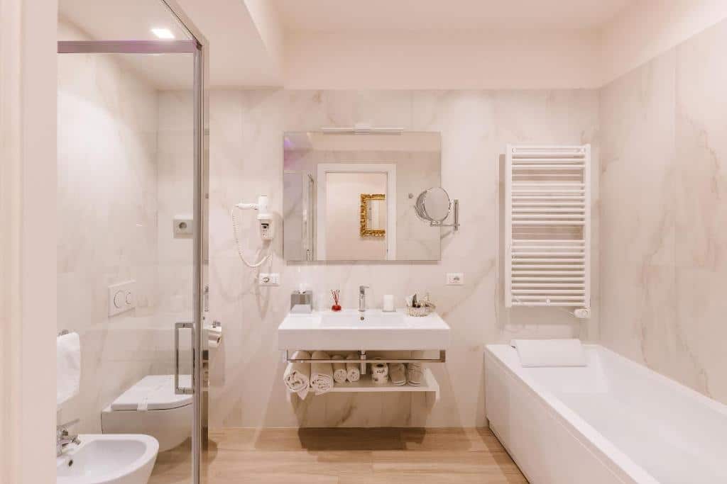 banheiro do Rhea Silvia Luxury Navona com banheira de hidro, box de vidro, pia com espaço embaixo, espelho e chuveirinho em tons de branco e dourado