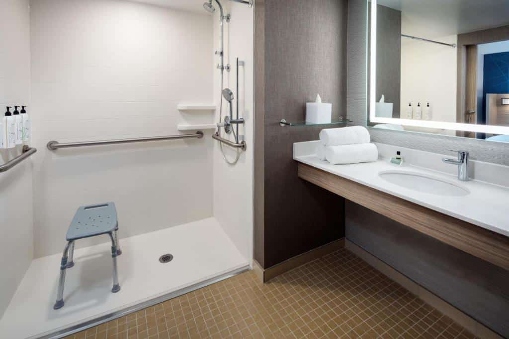 baheiro acessível do hotel SpringHill Suites by Marriott perto do aeroporto jfk em nova york com barras de apoio e uma cadeira de banho dentro do box, e uma pia vazada e ampla do lado direito.